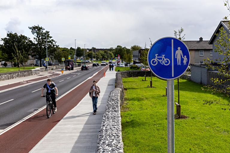 A photo from the Ballybane Road and Castlepark Road Cycle Network Scheme, showing a walker and a cyclist near to a blue sign